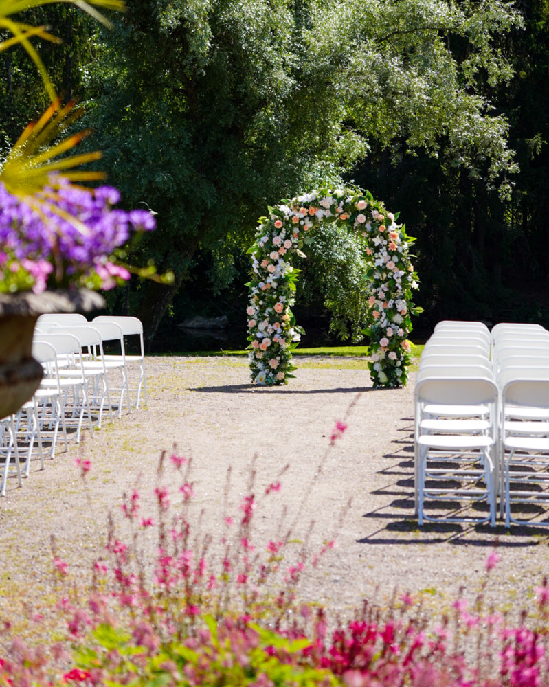 Outdoor wedding arch decorated with silk flowers, framed by rows of white chairs and flowering planters