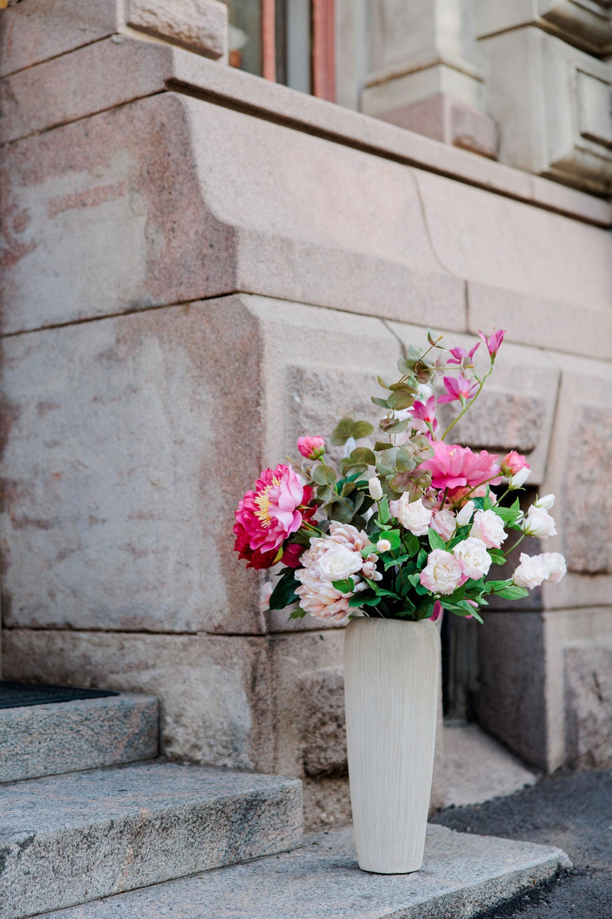 Runsas vaaleanpunainen ja valkoinen kukka-asetelma korkeassa vaaleassa maljakossa portailla.