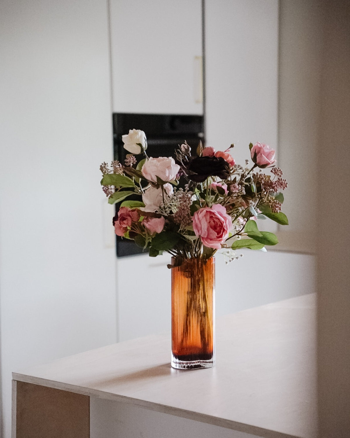 Violetta-silkkikukkakimppu aseteltuna korkeaan meripihkanväriseen maljakkoon.